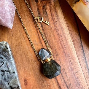 May include: A gold chain necklace featuring a dark grey, faceted gemstone pendant in a hexagonal bottle shape with a gold cap. A small gold key charm is near the clasp. The necklace is displayed on a wooden surface.