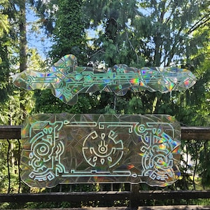 May include: Two clear, geometric-shaped decorative pieces with iridescent accents. The top piece is a key shape, and the bottom piece is a rectangle with a circular design. Both are displayed outdoors against a backdrop of trees.