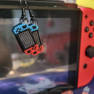 Puede incluir: Pendientes hechos a mano con un diseño pixelado que se asemeja a un mando de videojuegos. Los pendientes tienen ganchos negros y son principalmente negros, con detalles en azul y rojo. Cuelgan delante de una consola de juegos roja y negra.