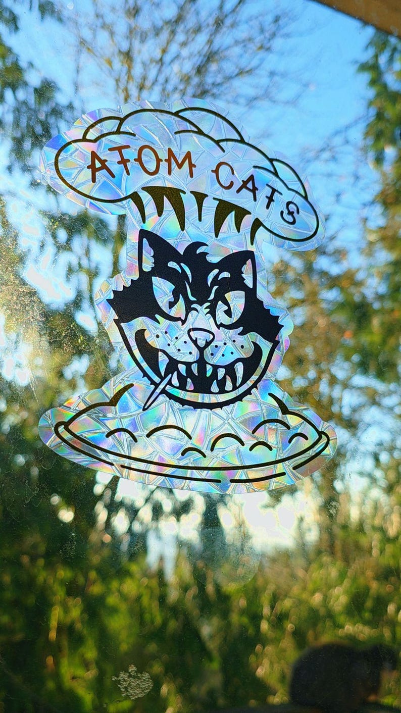 May include: Holographic sticker featuring a black cat with a menacing expression, a cigarette, and the text "ATOM CATS" above. The cat is on a UFO-shaped design. The sticker has a rainbow iridescent effect.
