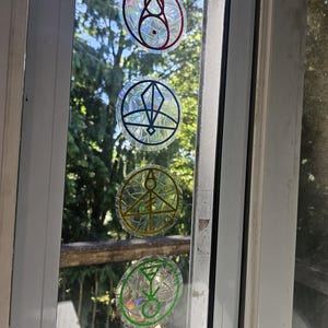 May include: Four circular holographic stickers with colorful geometric designs are adhered to a window. The stickers feature red, blue, yellow, and green outlines, with intricate patterns inside. The window frame is white, and a green tree is visible outside.