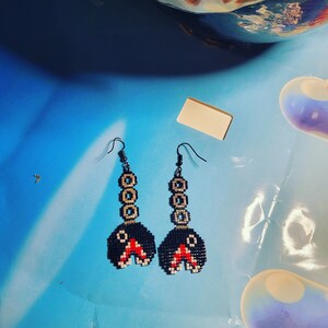 Pode incluir: Um par de brincos feitos à mão com um design pixelizado. Os brincos têm uma base preta com detalhes em vermelho e branco e uma série de círculos dourados. Os brincos estão pendurados em uma superfície azul.