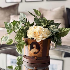 May include: A cylindrical brown planter with vertical stripes and a central oval portrait of a person in period clothing. The planter sits on a circular base and contains a cluster of cream-coloured artificial roses and green variegated foliage.