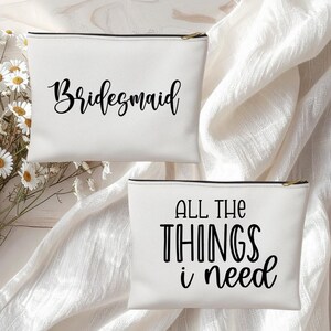 May include: Two white zippered cosmetic bags. One bag has the word "Bridesmaid" in black script. The other bag has the phrase "ALL THE THINGS i need" in black block letters and script. Both bags have black zippers.