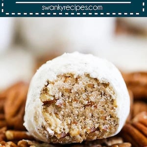 May include: A pecan snowball cookie, dusted with powdered sugar, is shown with a bite taken out, revealing the pecan filling. The cookie is sitting on a bed of pecan halves.