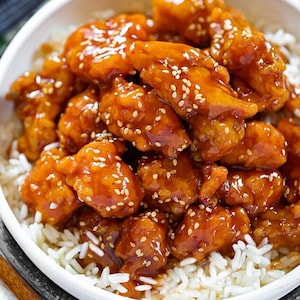May include: A white bowl filled with white rice topped with orange chicken glazed with a sweet and savory sauce and sprinkled with sesame seeds.