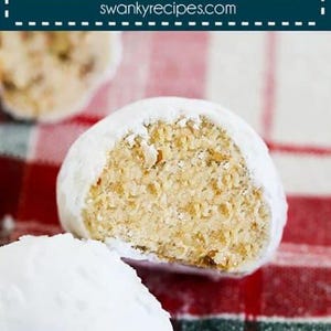 May include: Close-up of pecan snowballs, a type of cookie, on a red and green plaid tablecloth. The cookies are covered in powdered sugar and one is broken in half, revealing the pecan filling.