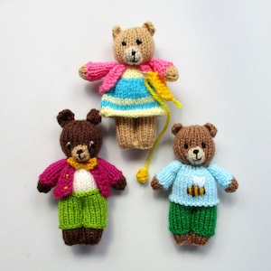 May include: Three knitted teddy bears wearing different outfits. The bear on the left is wearing a pink jacket, white shirt, and green pants. The bear in the middle is wearing a pink cardigan, blue and yellow striped dress, and has a yellow yarn tail. The bear on the right is wearing a blue sweater with a bee design and green pants.