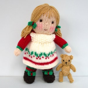 May include: A knitted doll with blonde hair, wearing a red and white dress with green accents, and brown shoes. The doll is holding a small brown knitted teddy bear.