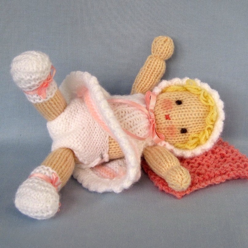 May include: A knitted doll with a pink dress, yellow hair, and a pink blanket. The doll is lying on its back with its arms outstretched.