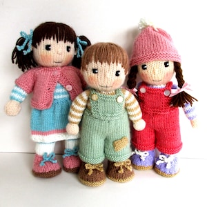 Puede incluir: Tres muñecas tejidas a mano. La muñeca de la izquierda lleva una rebeca rosa, un vestido azul y zapatos rosas. La muñeca del medio lleva un peto verde y zapatos marrones. La muñeca de la derecha lleva un peto rojo y un gorro rosa.