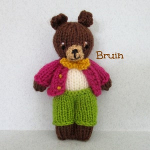May include: A brown knitted teddy bear wearing a pink cardigan with yellow buttons, a white shirt, a yellow bow tie, and green pants. The bear is standing on a white background. The text "Bruin" is written in brown letters on the right side of the image.