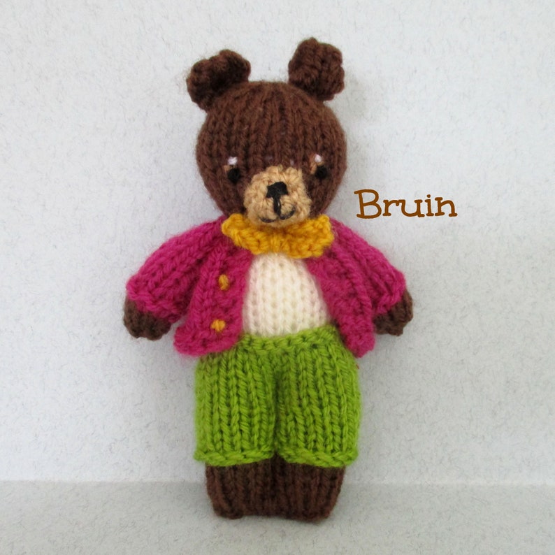 May include: A brown knitted teddy bear wearing a pink cardigan with yellow buttons, a white shirt, a yellow bow tie, and green pants. The bear is standing on a white background. The text "Bruin" is written in brown letters on the right side of the image.