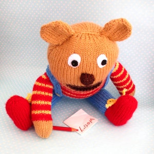 May include: A knitted brown bear wearing blue overalls with red and yellow stripes. The bear is sitting on a blue background with white polka dots. The bear is holding a red pencil and a note that says "worry".