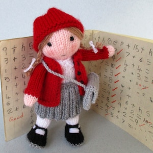 May include: A knitted doll wearing a red hooded sweater, a grey skirt, and black shoes. The doll is holding a grey knitted bag and is standing next to a notebook with handwritten math problems.