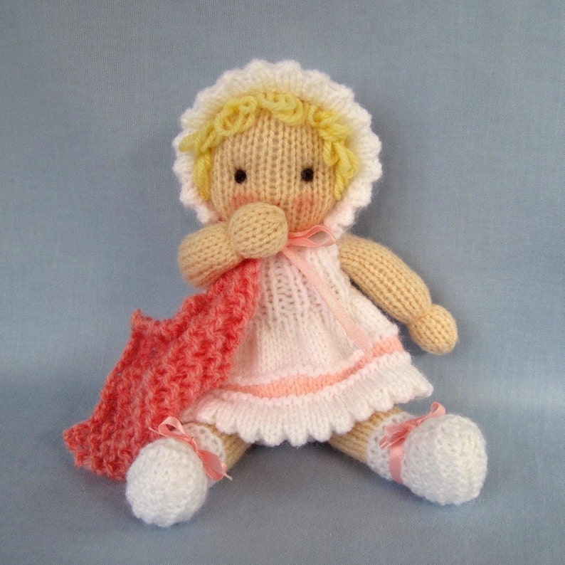 May include: A knitted doll with blonde hair, wearing a white dress with pink trim and a pink knitted blanket. The doll is sitting on a blue background.