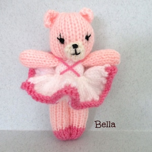 May include: A pink knitted ballerina bear wearing a white and pink tutu. The bear has black eyes and a black nose. The bear is labeled "Bella".