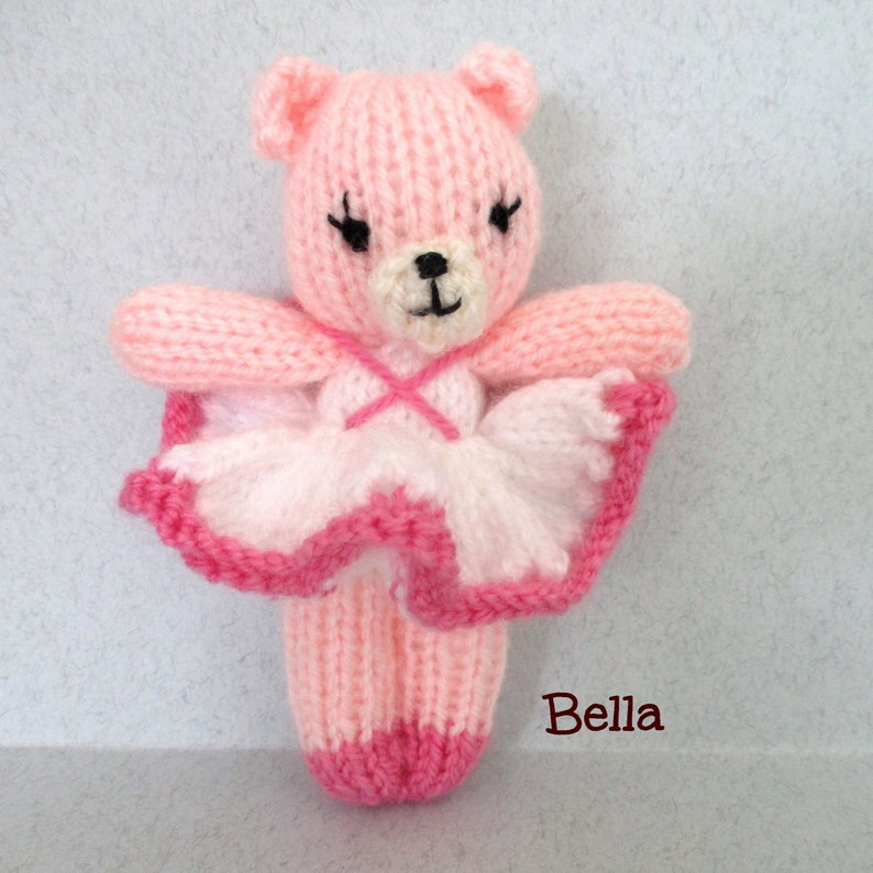 May include: A pink knitted ballerina bear wearing a white and pink tutu. The bear has black eyes and a black nose. The bear is labeled "Bella".