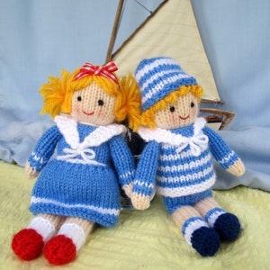 May include: Two knitted dolls dressed in blue and white sailor outfits. The girl doll has a red bow in her hair and the boy doll has a blue and white striped hat.