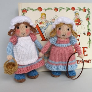 Puede incluir: Dos muñecas de punto vestidas con vestidos rosas y azules con gorros blancos. La muñeca de la izquierda lleva una cesta marrón y la muñeca de la derecha lleva un aro marrón.