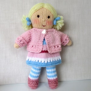 May include: A knitted doll with blonde hair, wearing a pink cardigan, a blue and white striped dress, and blue and white striped leggings. The doll has a white heart button on its cardigan.
