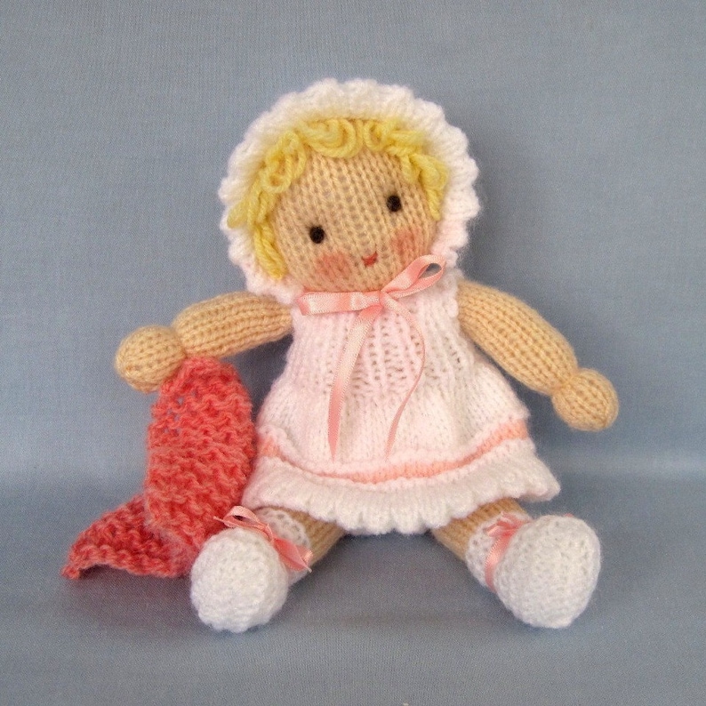 May include: A knitted doll with blonde hair, wearing a pink and white dress and a pink ribbon. The doll is holding a pink knitted blanket.