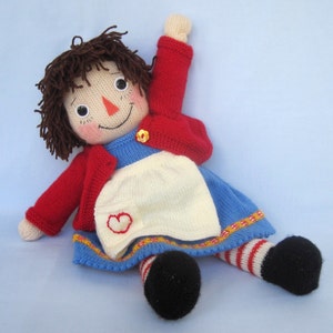 May include: A knitted rag doll with brown yarn hair, a red cardigan, a blue dress with a white apron, and red and white striped stockings. The doll is sitting on a light blue background.