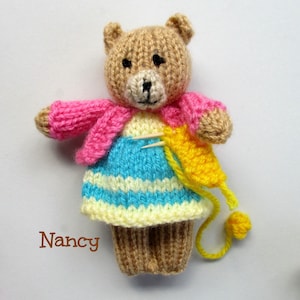 May include: A knitted teddy bear wearing a pink cardigan and a blue and yellow striped dress. The bear is holding a yellow yarn ball and a knitting needle.