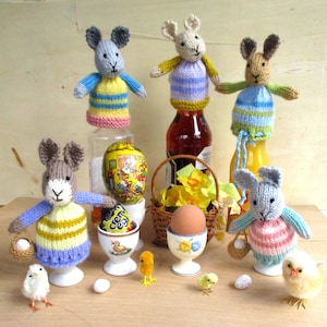 May include: A collection of handmade Easter decorations. The image features several knitted bunny figures, some perched on bottles, and others holding baskets. Also included are decorated eggs, chicks, and an egg in a decorative egg cup. The color palette includes pastels and natural tones.