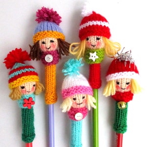 May include: A collection of handmade, knitted pen toppers in the shape of dolls. Each doll has a unique hat, scarf, and button details. The pens are in various colors, including red, yellow, blue, and green, with a variety of pom-poms and embellishments.