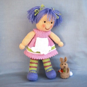 May include: A knitted doll with purple hair, a pink dress with a white apron, and green and brown striped leggings. The doll is holding a small knitted bunny with a carrot.