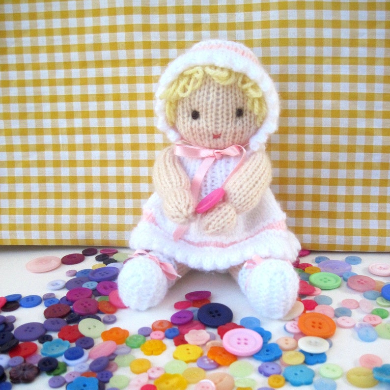 May include: A knitted doll with blonde hair, wearing a white and pink dress, sitting on a white surface surrounded by colorful buttons.