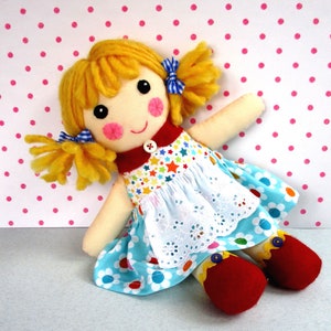 Puede incluir: Una muñeca de trapo hecha a mano con cabello de hilo amarillo peinado en coletas, adornada con lazos de cuadros vichy azules y blancos. La muñeca viste un vestido floral azul con un delantal de encaje blanco, un cuello rojo y zapatos rojos.