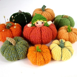 May include: A selection of hand-knitted pumpkins in shades of orange, green, and yellow. Some have green stalks, and one features a small doll wearing a green hat. The pumpkins are displayed on a white background.