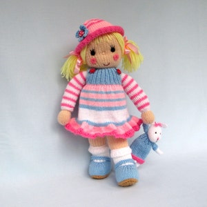 May include: A knitted doll with blonde hair, wearing a pink and blue striped dress, a pink hat with a flower, and blue shoes. The doll is holding a small knitted bunny.