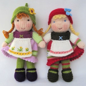 May include: Two knitted dolls wearing traditional German dirndl dresses. The doll on the left has green and purple clothing with a white apron and yellow flowers. The doll on the right has red and green clothing with a white apron and a blue flower.