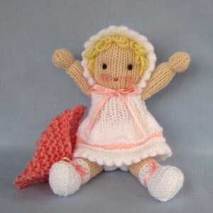May include: A knitted doll with blonde hair, wearing a white dress with pink trim and a pink ribbon. The doll is holding its arms out and has a pink knitted blanket beside it.