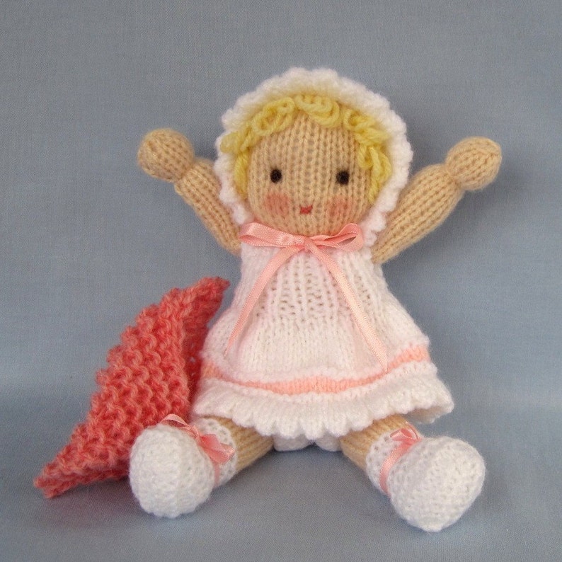 May include: A knitted doll with blonde hair, wearing a white dress with pink trim and a pink ribbon. The doll is holding its arms out and has a pink knitted blanket beside it.