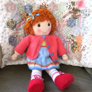 May include: A knitted doll with red hair, a pink cardigan, a blue dress, and red shoes. The doll is sitting on a patchwork quilt.