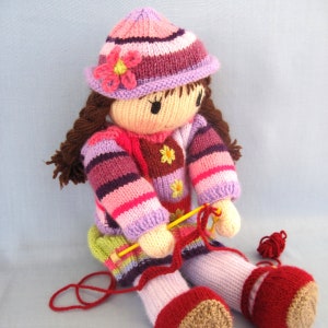 May include: A knitted doll wearing a pink and purple striped hat with a flower on top, a pink and purple striped jumper, and pink and purple striped trousers. The doll is holding a yellow knitting needle and a ball of red wool.
