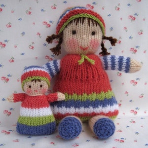 Puede incluir: Dos muñecas de punto, una más grande que la otra, ambas con vestidos y sombreros a rayas de colores. La muñeca más grande tiene el pelo castaño y un vestido rojo con rayas verdes y blancas. La muñeca más pequeña tiene un vestido a rayas rosa y blanco con rayas verdes y azules.