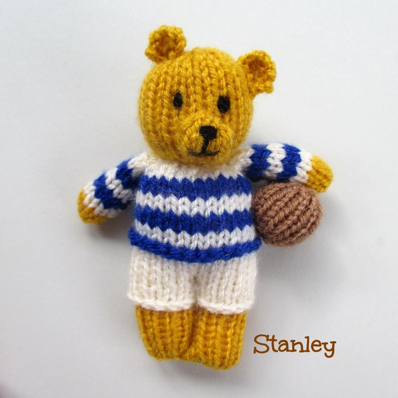 May include: A yellow knitted teddy bear wearing a blue and white striped shirt and white shorts. The bear is holding a brown ball and the text "Stanley" is written below the bear.