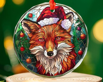 May include: A round, clear Christmas ornament featuring a fox wearing a Santa hat. The fox is depicted in shades of orange, red, and white, with a detailed stained-glass style design. Red ribbon for hanging. Ornaments and greenery are visible.