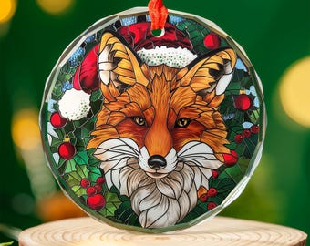 May include: A round, clear glass Christmas ornament featuring a fox wearing a Santa hat. The fox has orange and white fur, with a detailed stained-glass style design. Red berries and green leaves surround the fox, with a red ribbon for hanging.