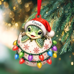 May include: A hanging Christmas ornament featuring a green baby turtle hatching from a white eggshell decorated with red snowflakes. The turtle wears a red Santa hat with the name Emma printed on the brim. A string of colorful holiday lights wraps around the egg.