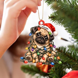 May include: A flat, decorative Christmas ornament featuring a tan pug puppy wearing a red Santa hat with the name Lucas printed on the white trim. The pug is wrapped in a string of colorful, glowing Christmas lights in red, blue, green, and yellow.