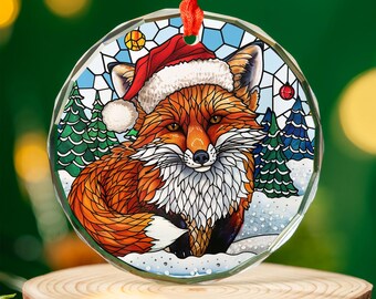 May include: A round Christmas ornament featuring a fox wearing a Santa hat. The fox has orange fur and white accents, set against a winter scene with snow and pine trees. The ornament hangs from a red ribbon.