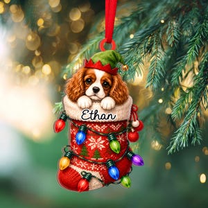 May include: A flat Christmas ornament featuring a Cavalier King Charles Spaniel puppy wearing a green elf hat, peeking out of a red and white knit stocking. The stocking is decorated with a string of colorful lights and the name Ethan.