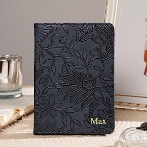 Può includere: Un diario in pelle blu scuro con un motivo floreale in rilievo. Il diario presenta il nome "Max" in lettere dorate. È esposto su una superficie beige, insieme a una foto incorniciata e un portacandele in vetro.