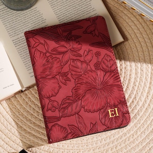 May include: A burgundy passport holder with a floral design and gold initials "EI". The cover is embossed with a detailed pattern of hibiscus flowers and leaves. The holder is resting on a woven mat, next to a candle and a perfume bottle.